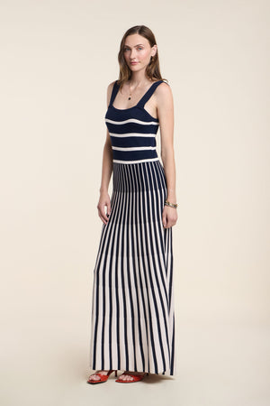 A woman wears the AVELINE DRESS by Velvet by Graham & Spencer, a sleeveless knit maxi with a fitted bodice, navy and white horizontal stripes on top, vertical stripes on the skirt, styled with red sandals.