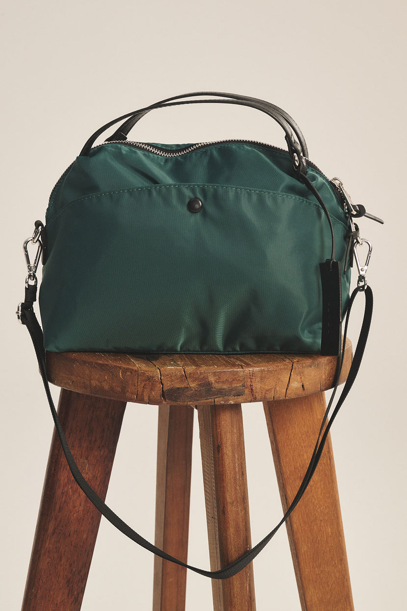 The NYLON CROSSBODY BAG by Velvet by Graham & Spencer, crafted from durable green nylon with black handles and a removable shoulder strap, is displayed on a wooden stool against a plain background.