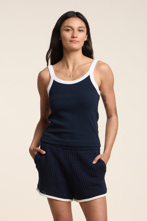 A woman wears the BELLE TANK TOP by Velvet by Graham & Spencer, a navy ribbed tank with a scoop neckline and white trim, paired with matching textured shorts, standing hands in pockets against a plain backdrop.