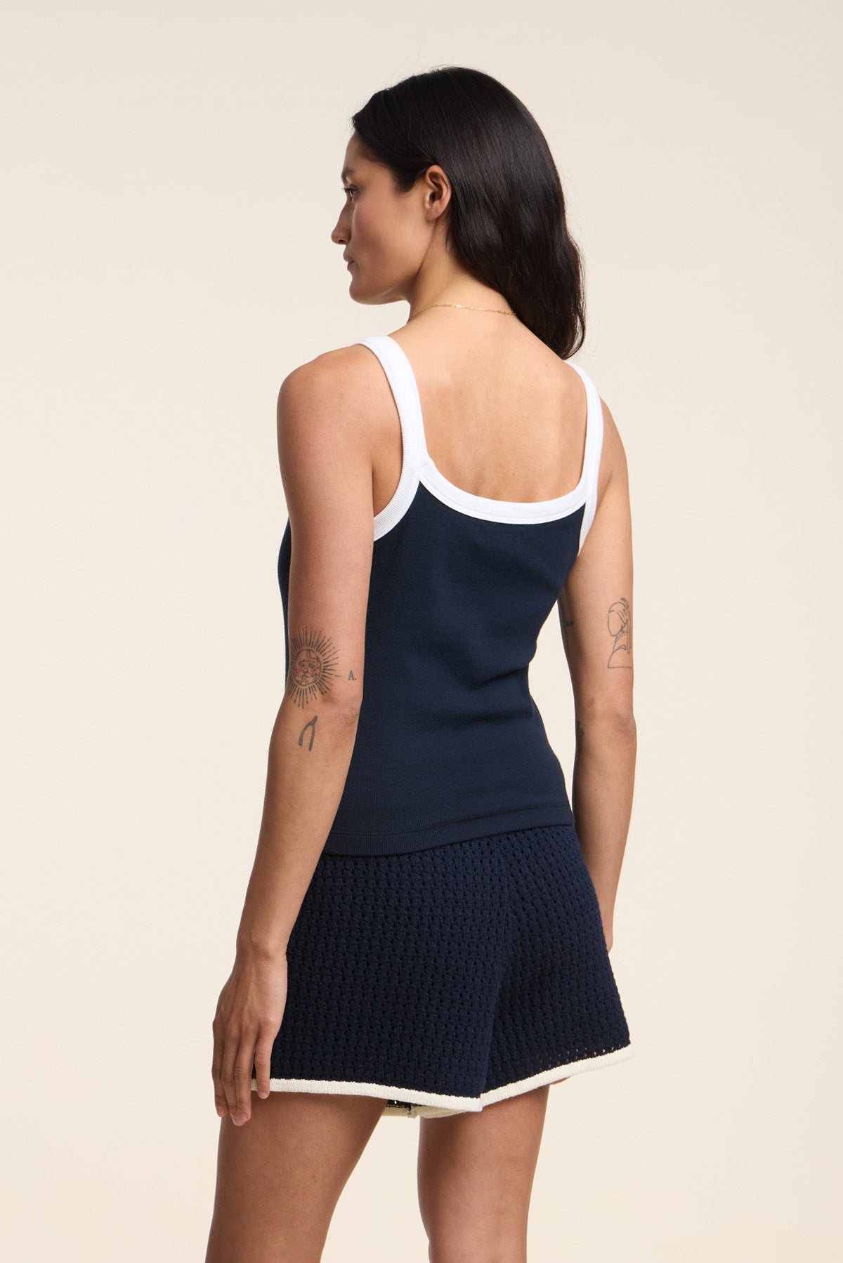 A woman faces away, wearing the BELLE TANK TOP by Velvet by Graham & Spencer—a navy ribbed tank with white straps—paired with navy shorts trimmed in white. The scoop neckline and her arm tattoos add stylish details.