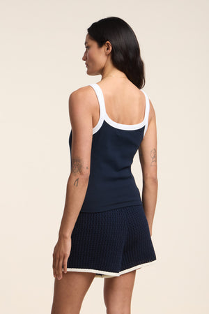 A woman faces away, wearing the BELLE TANK TOP by Velvet by Graham & Spencer—a navy ribbed tank with white straps—paired with navy shorts trimmed in white. The scoop neckline and her arm tattoos add stylish details.