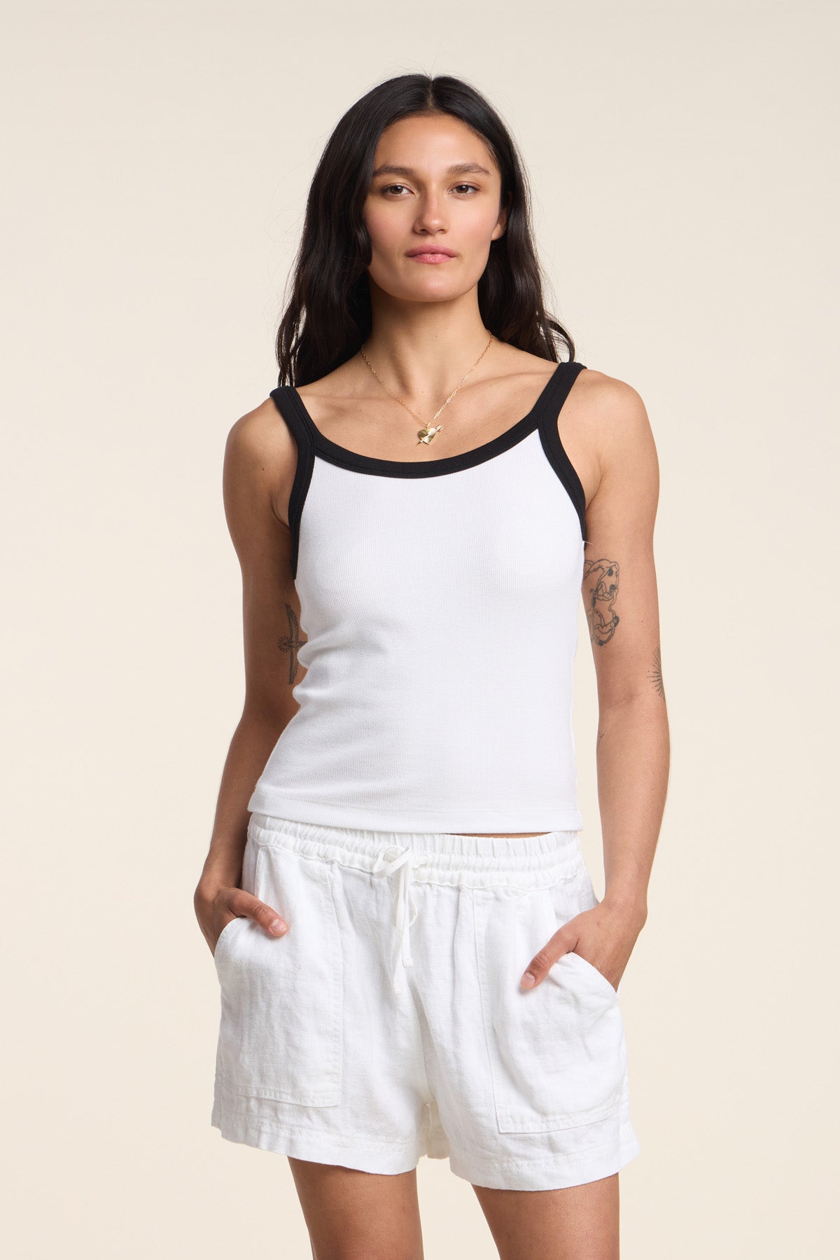 A woman stands with her hands in her pockets, wearing the BELLE TANK TOP by Velvet by Graham & Spencer—featuring a white ribbed design with black trim—paired with white shorts against a plain background.-41644968378561
