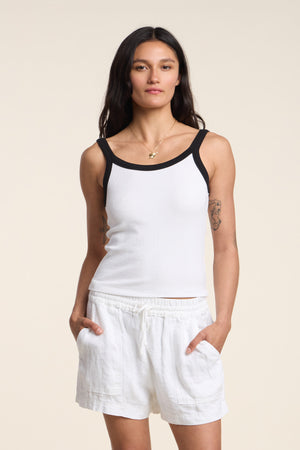 A woman stands with her hands in her pockets, wearing the BELLE TANK TOP by Velvet by Graham & Spencer—featuring a white ribbed design with black trim—paired with white shorts against a plain background.