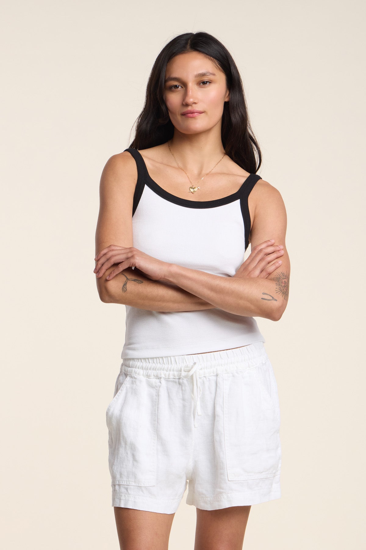 A woman with long dark hair stands with arms crossed, wearing the BELLE TANK TOP by Velvet by Graham & Spencer—a ribbed scoop-neck tank with black trim—paired with white drawstring shorts, against a plain light background.