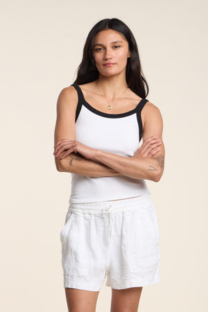 A woman with long dark hair stands with arms crossed, wearing the BELLE TANK TOP by Velvet by Graham & Spencer—a ribbed scoop-neck tank with black trim—paired with white drawstring shorts, against a plain light background.