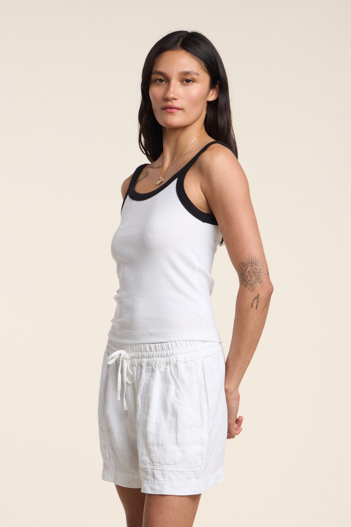 A woman with long dark hair wears the BELLE TANK TOP by Velvet by Graham & Spencer, featuring a ribbed texture, scoop neckline, and black trim. She pairs it with white drawstring shorts and has a sun tattoo on her upper left arm.