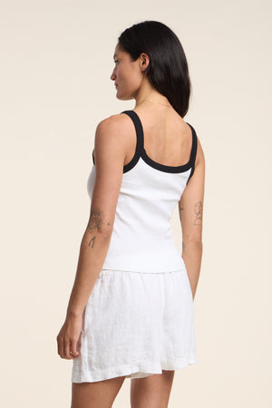A woman with visible tattoos stands sideways, wearing the BELLE TANK TOP by Velvet by Graham & Spencer—white, ribbed, with black trim and a scoop neckline—paired with a white skirt.