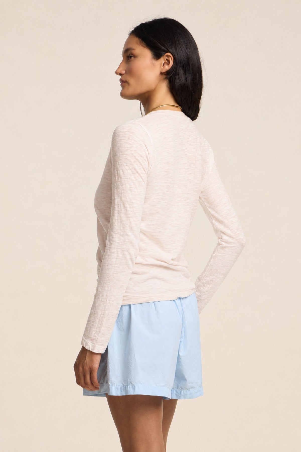 A woman stands sideways against a plain background, wearing the BLAIRE TEE by Velvet by Graham & Spencer—a textured cotton slub long sleeve tee with a v-neckline—paired with light blue shorts.-41775044559041