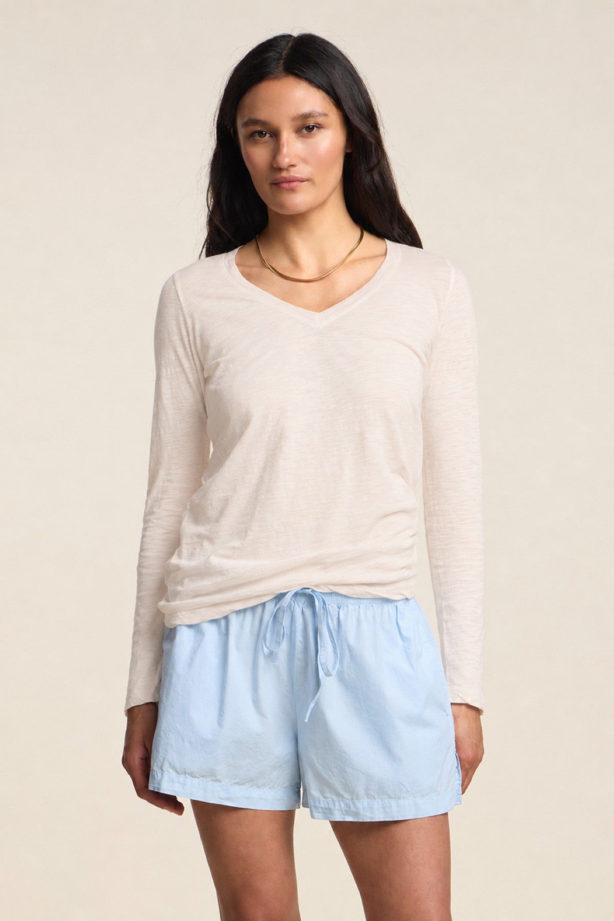 A woman stands against a plain background wearing the BLAIRE TEE by Velvet by Graham & Spencer—a beige long sleeve tee with a flattering v-neckline—paired with light blue drawstring shorts.