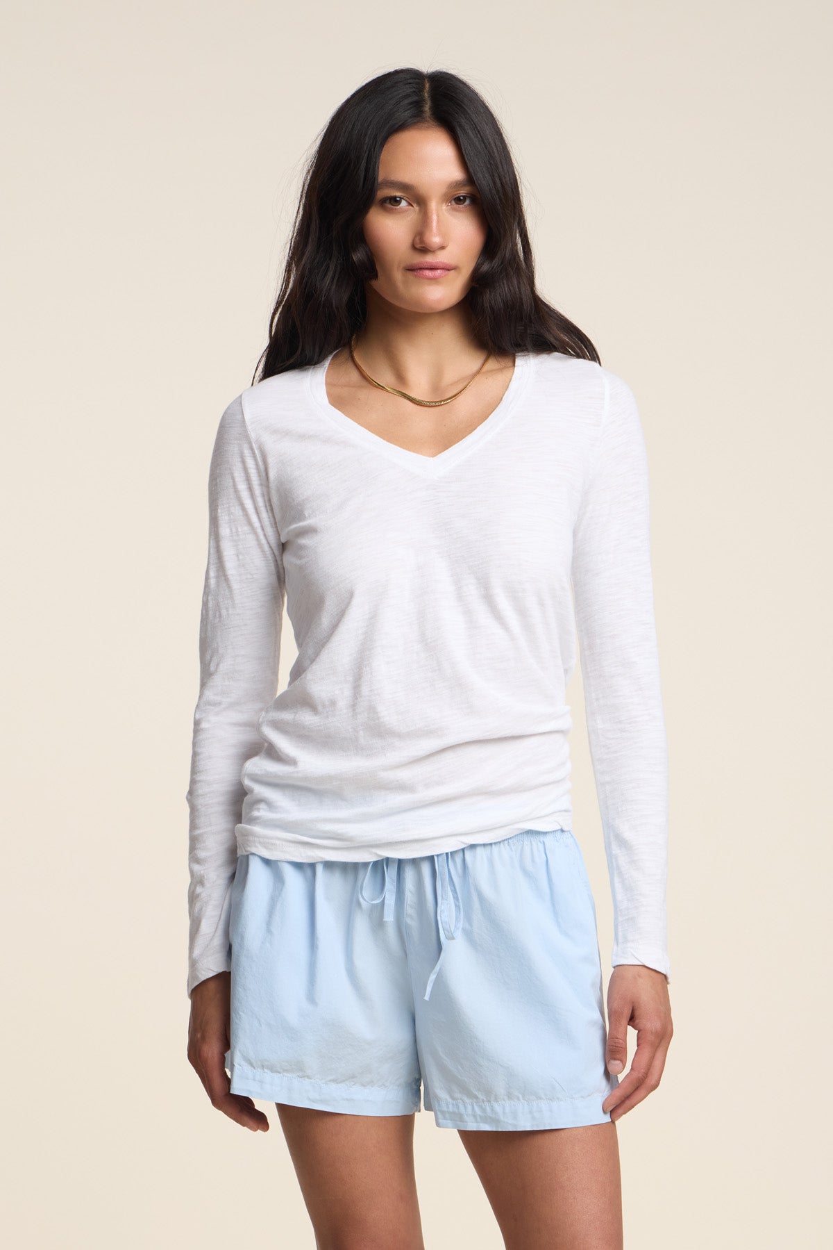 A woman with long dark hair stands facing the camera against a plain beige background, wearing the BLAIRE TEE by Velvet by Graham & Spencer—a white cotton slub top featuring a flattering V-neckline and long sleeves—paired with light blue drawstring shorts.