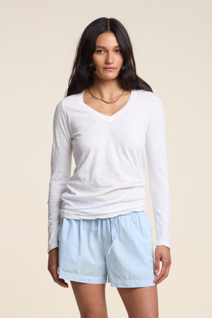 A woman with long dark hair stands facing the camera against a plain beige background, wearing the BLAIRE TEE by Velvet by Graham & Spencer—a white cotton slub top featuring a flattering V-neckline and long sleeves—paired with light blue drawstring shorts.