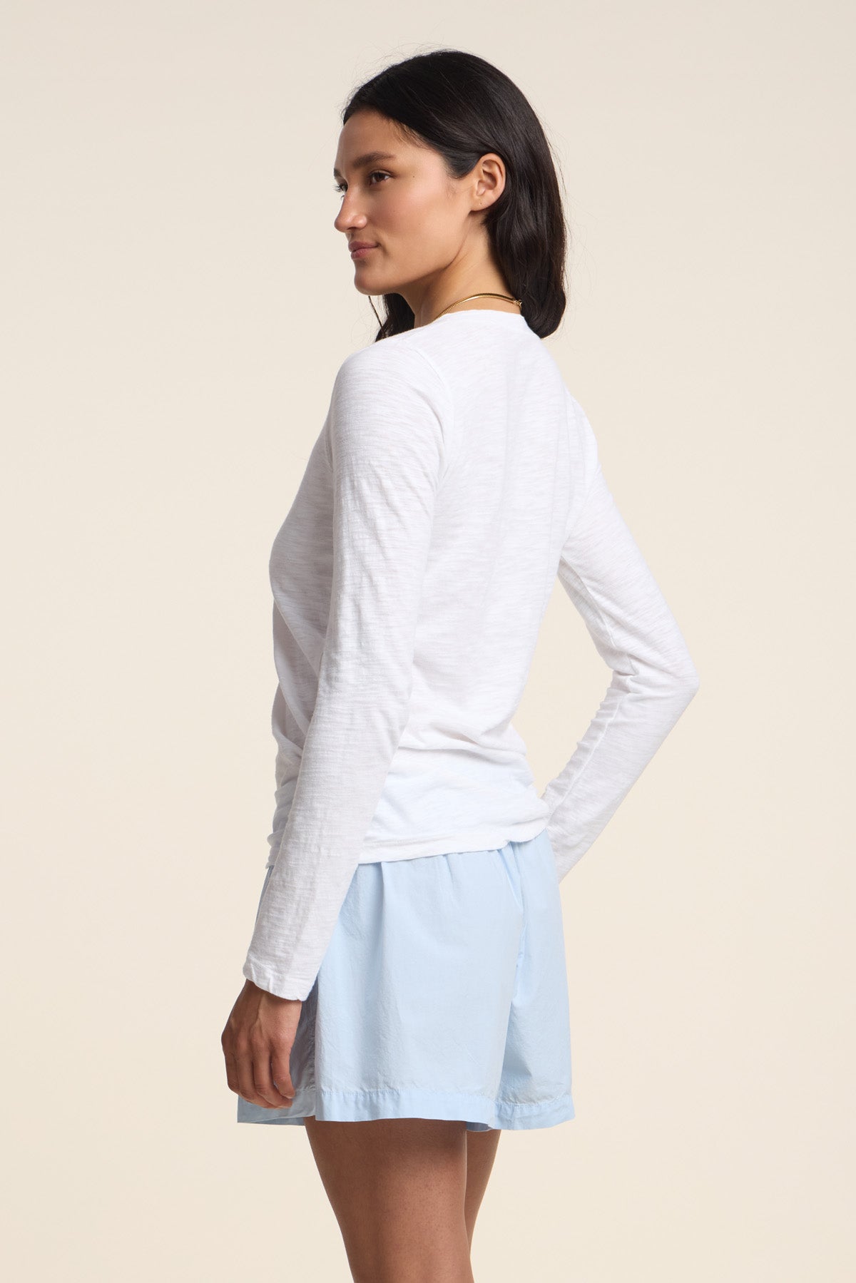 A woman stands sideways against a plain background, one hand on her hip, wearing the Velvet by Graham & Spencer BLAIRE TEE—a long-sleeve cotton slub top with a v-neckline—paired with light blue shorts.-41775118811329