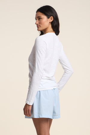 A woman stands sideways against a plain background, one hand on her hip, wearing the Velvet by Graham & Spencer BLAIRE TEE—a long-sleeve cotton slub top with a v-neckline—paired with light blue shorts.