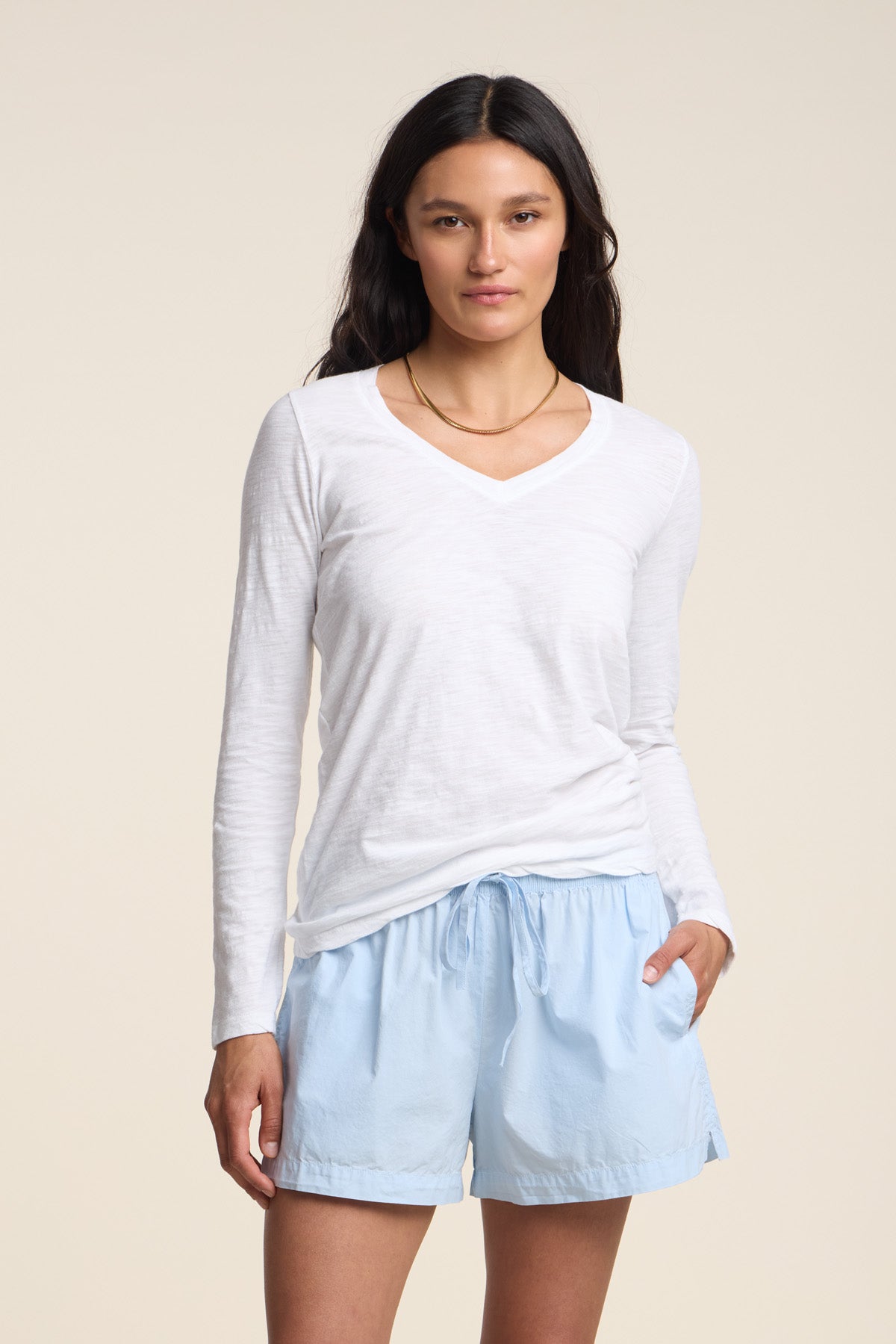 A woman with long dark hair wears the Velvet by Graham & Spencer BLAIRE TEE—a white cotton slub V-neckline long sleeve top—paired with light blue drawstring shorts, standing against a plain light background.
