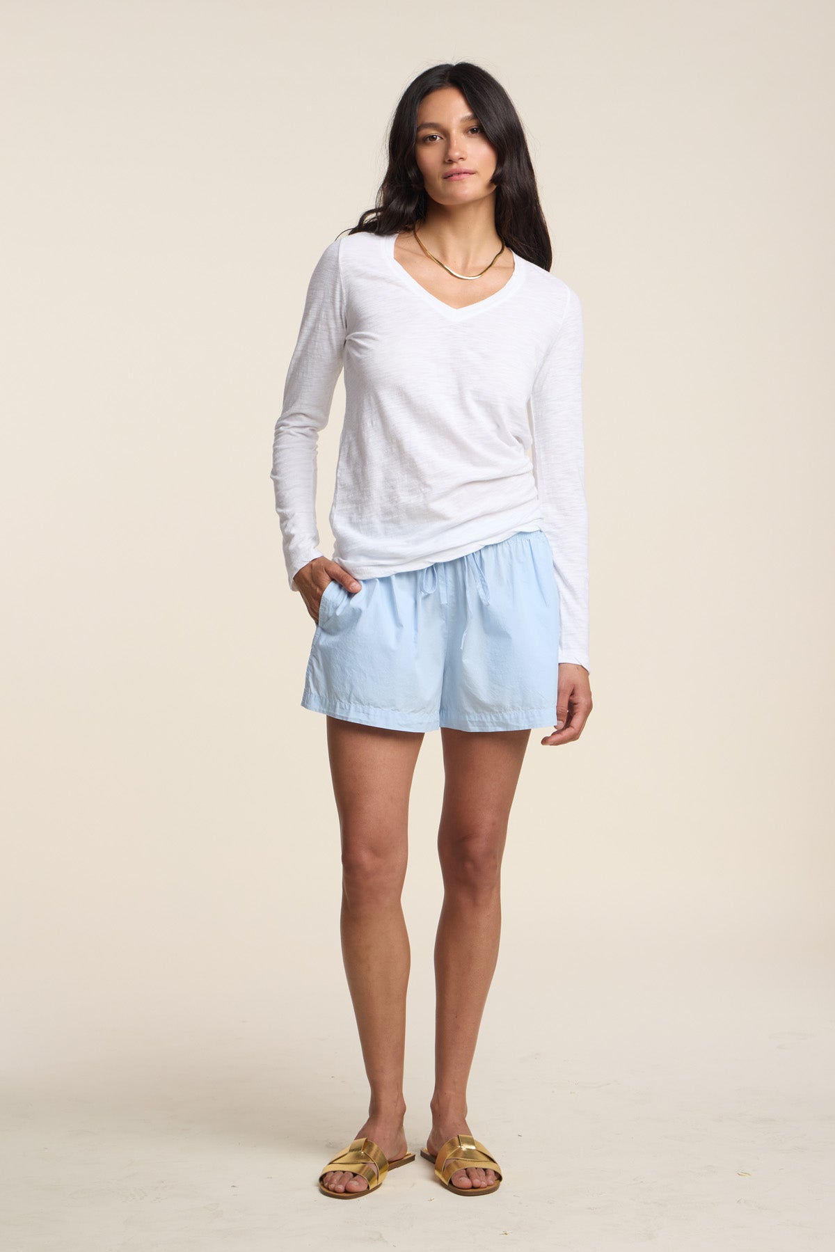 A woman stands facing forward against a plain background, wearing the BLAIRE TEE by Velvet by Graham & Spencer—a white cotton slub top featuring long sleeves and a subtle v-neckline—paired with light blue shorts and gold sandals, with one hand in her pocket.