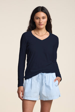 A woman stands against a plain background wearing the Velvet by Graham & Spencer BLAIRE TEE, a navy cotton slub long sleeve v-neck, paired with light blue drawstring shorts and one hand in her pocket.