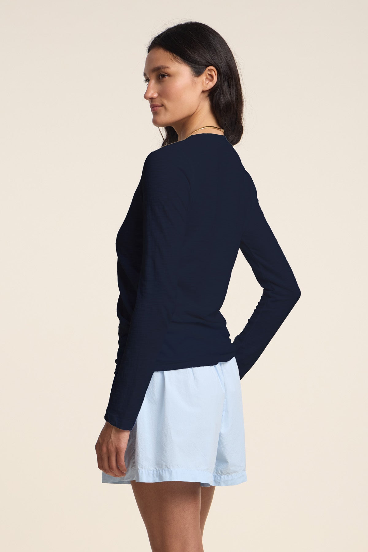 A woman stands in a side-facing pose wearing the Velvet by Graham & Spencer BLAIRE TEE, a long sleeve cotton slub v-neck top, paired with light blue shorts against a plain background.