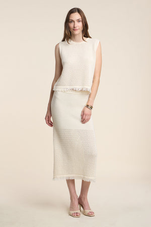 A woman wears the BLYTHE SKIRT by Velvet by Graham & Spencer, a cream cotton mesh pencil skirt with fringe hem, paired with a sleeveless knit top and neutral heels, standing against a beige background.