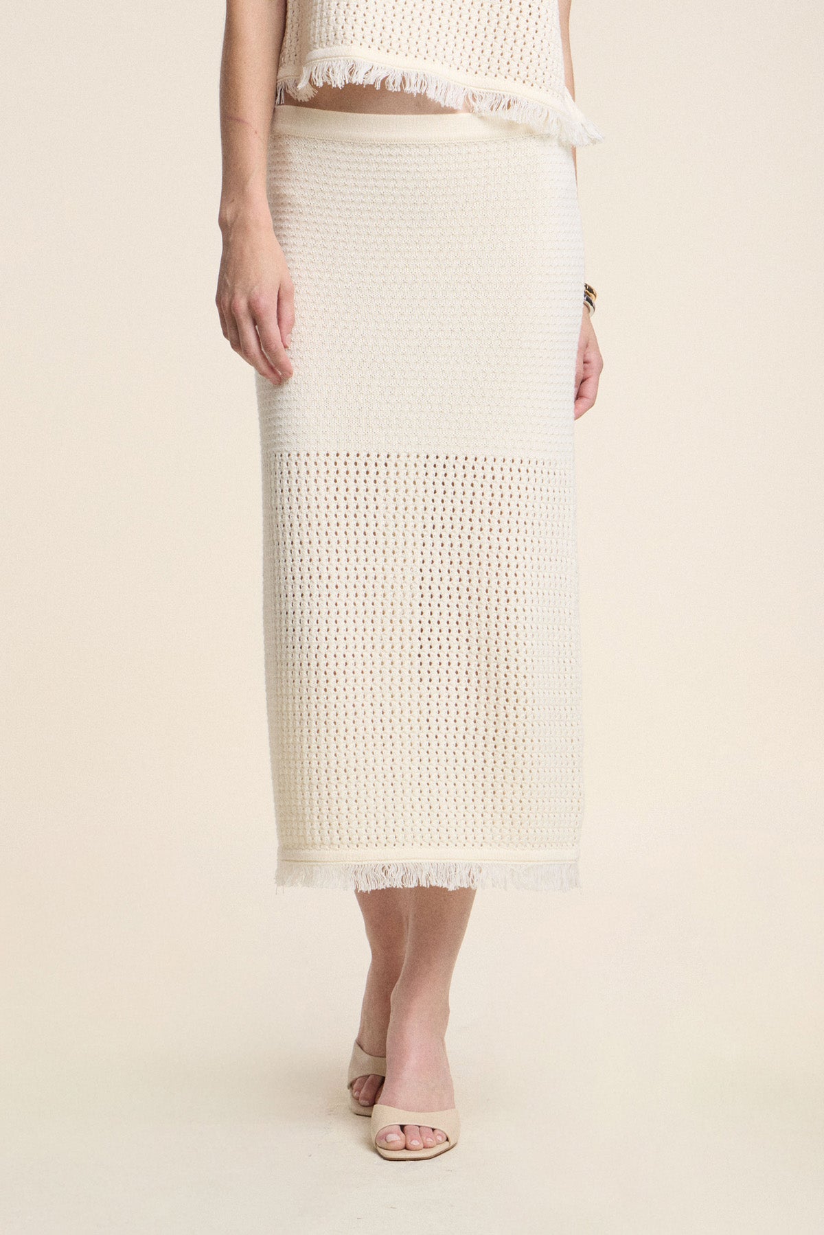A person wears the BLYTHE SKIRT by Velvet by Graham & Spencer, a cream ankle-length cotton mesh skirt with a fringe hem, paired with a matching top and beige open-toe heels.