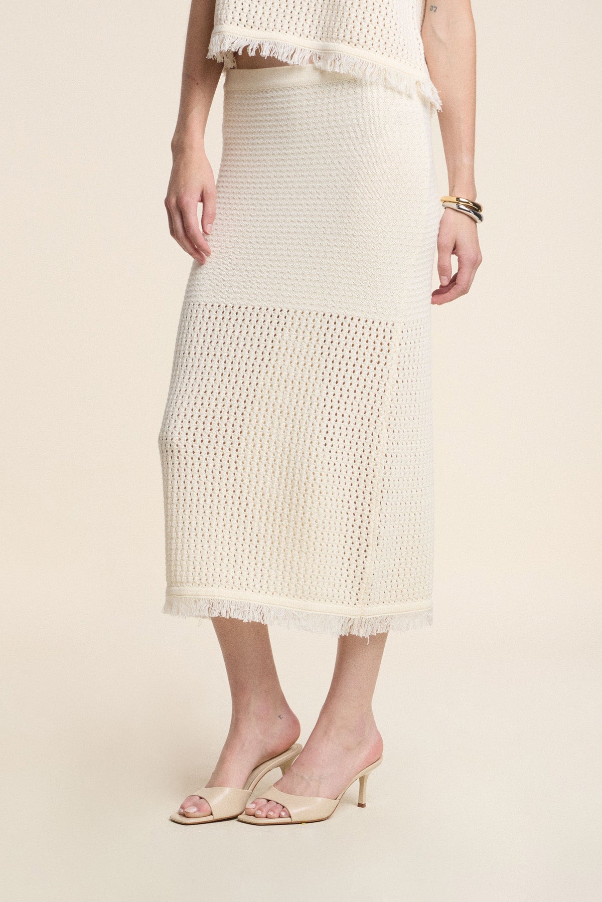 A person wearing the BLYTHE SKIRT by Velvet by Graham & Spencer—a cream-colored cotton mesh pencil skirt with a fringe hem—paired with nude heeled sandals and gold bracelets.