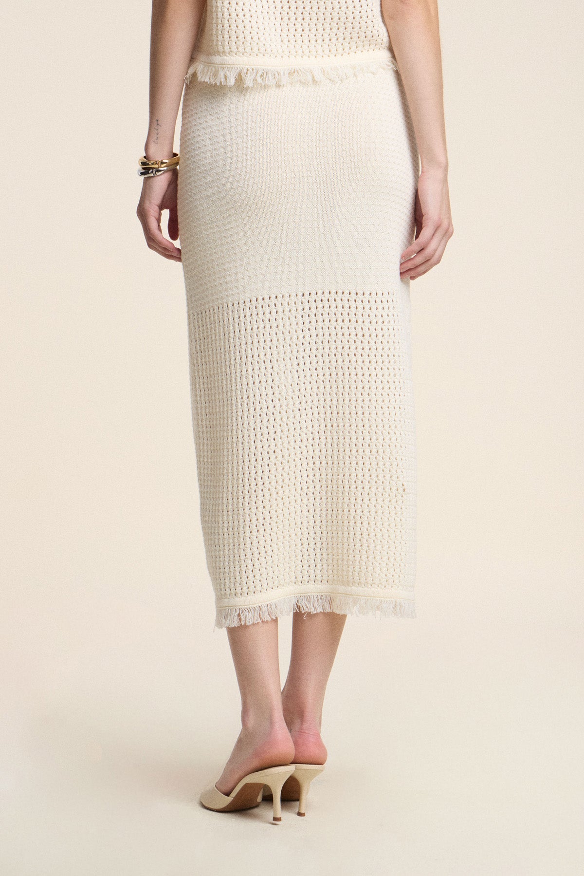 A person wears the BLYTHE SKIRT by Velvet by Graham & Spencer, a cream cotton mesh pencil skirt with fringe hem, styled with nude heeled mules and bracelets, photographed from the back against a plain background.-41374538956993