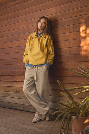 A woman wears the BOBBIE HOODIE in yellow by Velvet by Graham & Spencer, layered over a blue shirt and paired with beige oversized pants, as she stands against a wooden wall on a porch. A potted plant sits in the foreground.