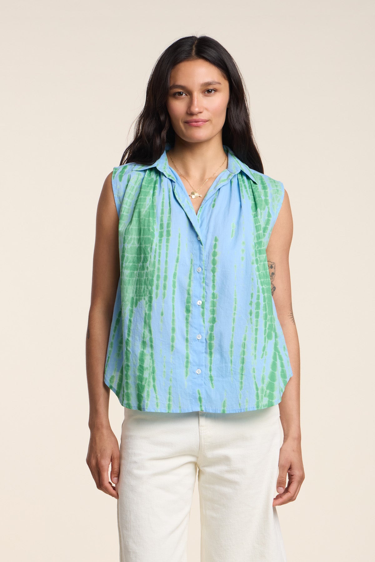 A woman stands facing the camera against a neutral background, wearing the Velvet by Graham & Spencer CALLISTO SHIRT—a relaxed fit, sleeveless blue and green tie-dye button-up—paired with white pants.-41644996264129