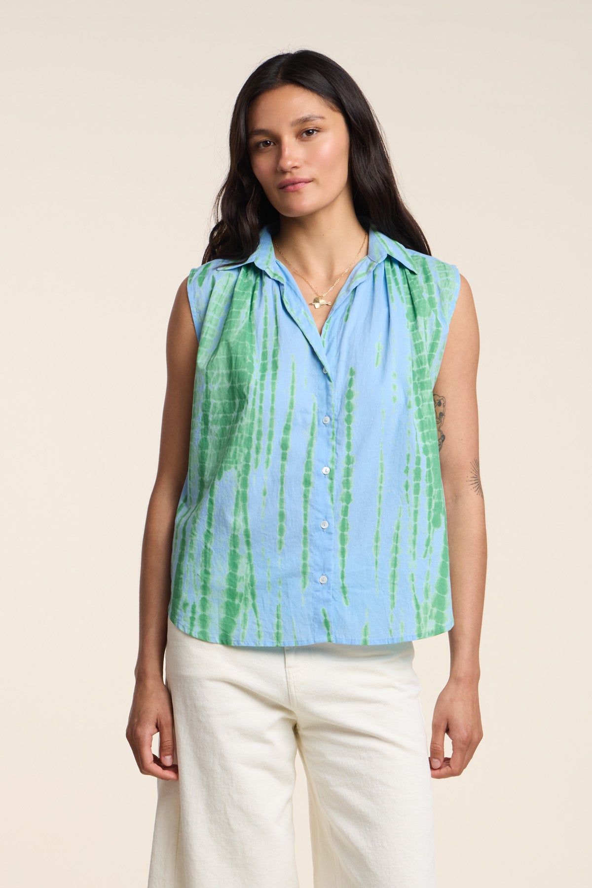 A woman models the CALLISTO SHIRT by Velvet by Graham & Spencer—a relaxed fit, sleeveless blue and green tie dye button-up—paired with white pants against a plain background.