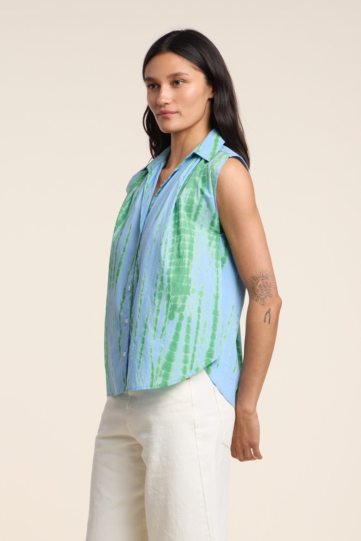 A woman wears the CALLISTO SHIRT by Velvet by Graham & Spencer, a relaxed fit, sleeveless blue and green tie dye button-up, paired with off-white pants. She has long dark hair and a circular tattoo on her upper arm.
