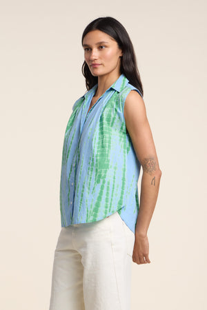 A woman wears the CALLISTO SHIRT by Velvet by Graham & Spencer, a relaxed fit, sleeveless blue and green tie dye button-up, paired with off-white pants. She has long dark hair and a circular tattoo on her upper arm.