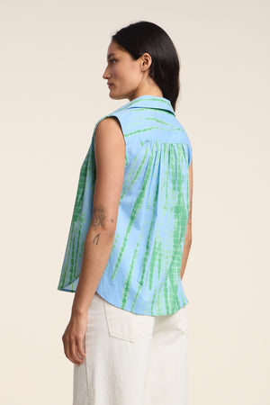 A woman stands in profile wearing the CALLISTO SHIRT by Velvet by Graham & Spencer, a blue and green sleeveless button-up with a relaxed fit, paired with white pants and displaying tattoos on her arm.