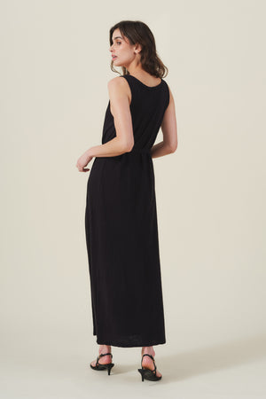 A woman stands facing away in the Velvet by Graham & Spencer CAMILA DRESS, a black cotton tank-style maxi, paired with black heeled sandals against a plain light background.