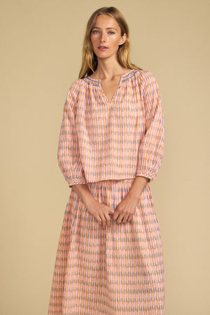 A woman stands against a beige background, wearing the CAMPBELL TOP by Velvet by Graham & Spencer—a loose-fitting top with a relaxed silhouette and pink, orange, and blue stripes.