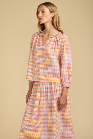 A woman poses against a beige background in the Velvet by Graham & Spencer CAMPBELL TOP, a relaxed-fit printed cotton V-neck paired with a matching pastel vertical striped skirt.