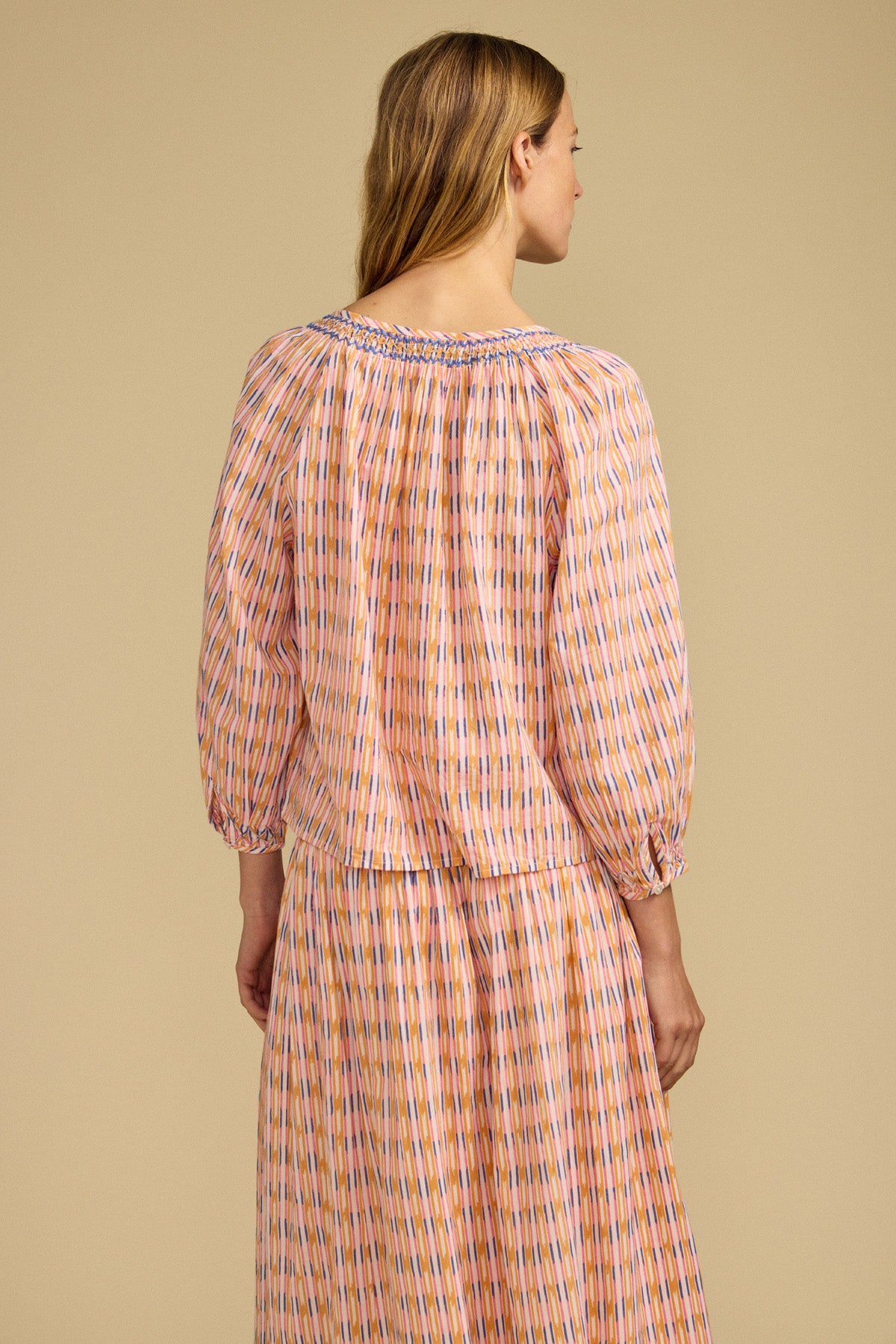 A woman with light brown hair is seen from the back wearing the CAMPBELL TOP by Velvet by Graham & Spencer, a relaxed-fit printed cotton top with long sleeves, paired with a matching skirt, set against a plain beige background.