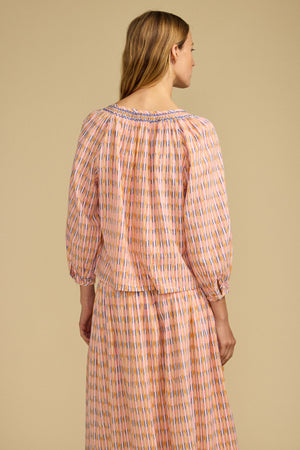 A woman with light brown hair is seen from the back wearing the CAMPBELL TOP by Velvet by Graham & Spencer, a relaxed-fit printed cotton top with long sleeves, paired with a matching skirt, set against a plain beige background.