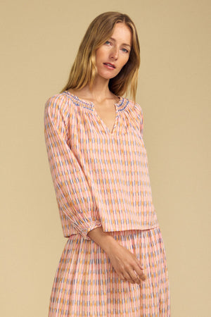 A woman wearing the CAMPBELL TOP by Velvet by Graham & Spencer, a relaxed silhouette, long-sleeved printed cotton piece, and a matching skirt poses against a beige background, looking at the camera.