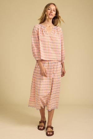 A woman stands against a beige background wearing Velvet by Graham & Spencer’s DEBORAH SKIRT, a printed cotton skirt with a smocked elastic waist, paired with a loose, long-sleeve pink and blue blouse and brown sandals.
