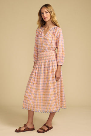 A woman stands in a studio wearing the CAMPBELL TOP by Velvet by Graham & Spencer, a long-sleeved, pink and orange printed cotton top with a V-neckline and smocked waist, paired with brown flat sandals.