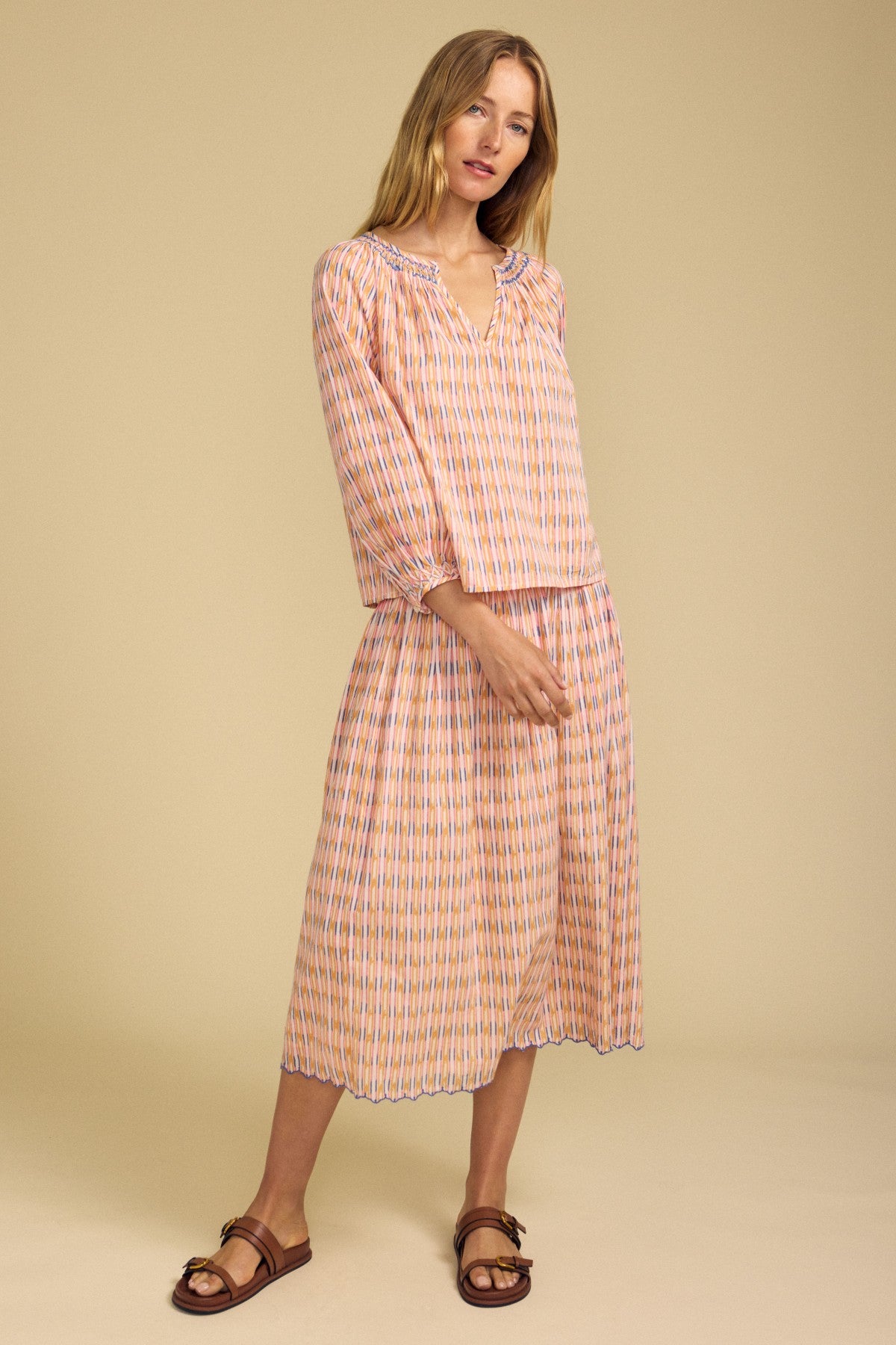 A woman stands against a beige background wearing a loose, long-sleeve patterned blouse and the DEBORAH SKIRT by Velvet by Graham & Spencer, featuring cotton stripes, a smocked elastic waist, and paired with flat sandals.