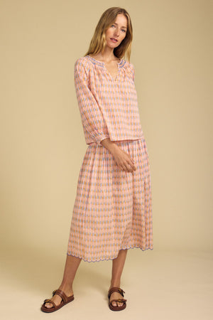 A woman stands against a beige background wearing a loose, long-sleeve patterned blouse and the DEBORAH SKIRT by Velvet by Graham & Spencer, featuring cotton stripes, a smocked elastic waist, and paired with flat sandals.