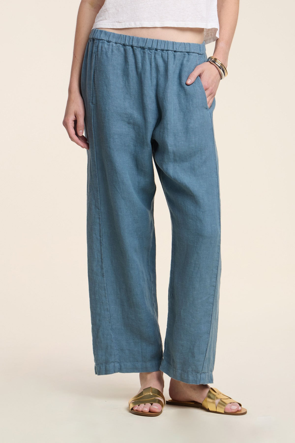 Wearing the CELINE HEAVY LINEN PANT by Velvet by Graham & Spencer—wide leg, cropped, blue with an elastic waist—styled with a white shirt, gold sandals, and bracelets; one hand in pocket against a plain background.-41371568537793