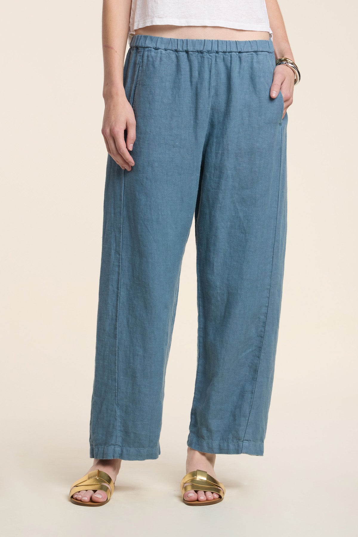 A person stands against a plain light background wearing Velvet by Graham & Spencer's CELINE HEAVY LINEN PANT—wide leg, cropped, elastic waist—paired with a white cropped top, gold sandals, and bracelets.