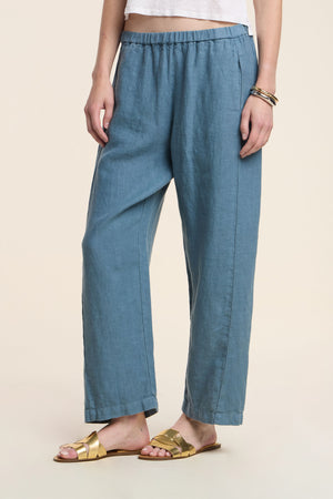 A person in Velvet by Graham & Spencer's CELINE HEAVY LINEN PANT, paired with a white top, gold sandals, and bracelets, stands against a neutral background.