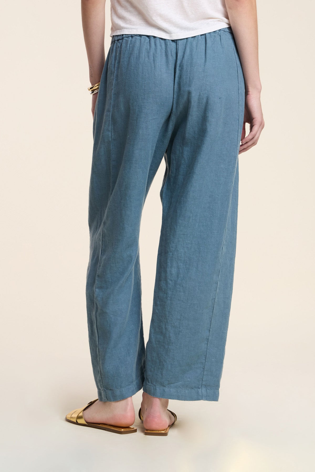 A person stands with their back to the camera, wearing Velvet by Graham & Spencer's CELINE HEAVY LINEN PANT in blue (wide-leg, cropped, elastic waist), a white top, gold bracelet, and gold open-toe flat sandals.