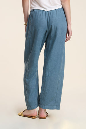 A person stands with their back to the camera, wearing Velvet by Graham & Spencer's CELINE HEAVY LINEN PANT in blue (wide-leg, cropped, elastic waist), a white top, gold bracelet, and gold open-toe flat sandals.