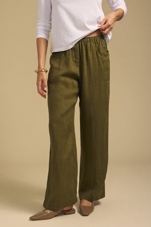 A person wearing the CLAIRO LINEN PANT by Velvet by Graham & Spencer in olive green—featuring a loose, wide-leg silhouette and elastic pull-on waist—paired with a white long-sleeve shirt, tan pointed flats, and a gold bracelet, stands against a plain beige background.