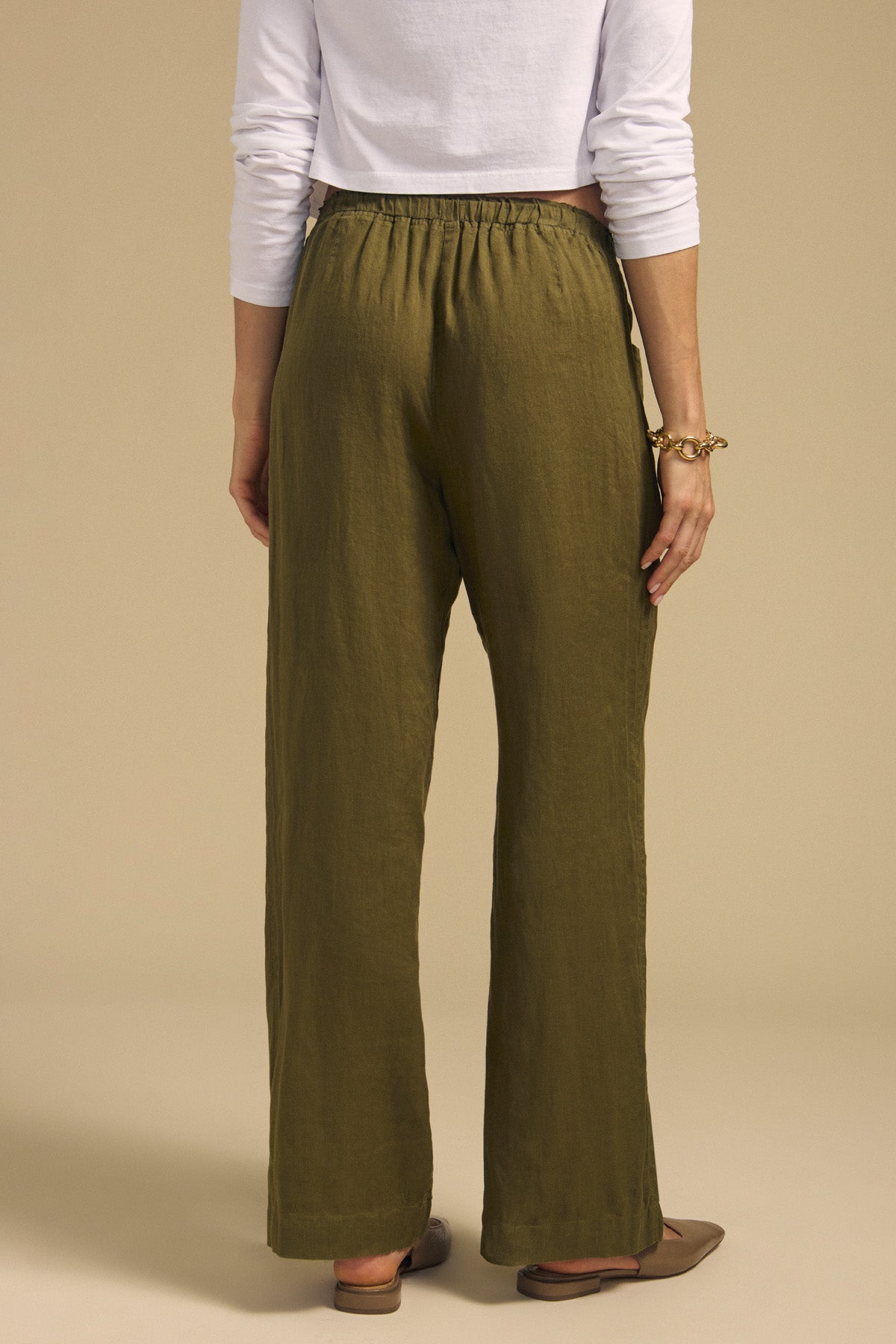 A person is shown from the back, standing against a neutral background, wearing the CLAIRO LINEN PANT by Velvet by Graham & Spencer in olive green—a lightweight linen pant with a wide-leg silhouette—paired with a white long-sleeve crop top, brown flats, and a gold bracelet.
