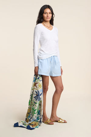 A woman wears the CLOVE SHORT by Velvet by Graham & Spencer with a white long-sleeve shirt, gold sandals, and holds a patterned scarf against a plain background.