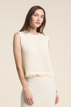 A woman wears the COVA SWEATER by Velvet by Graham & Spencer—a sleeveless, cream cotton knit top with fringe hem—paired with a matching skirt, standing against a beige background.